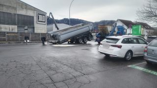 A white car drives past as a dump truck tipped over in the road. A man pours water on the truck. A crane is positioned behind the truck.