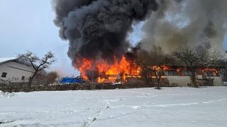 A building is engulfed in flames and smoke in a snowy area, surrounded by trees.