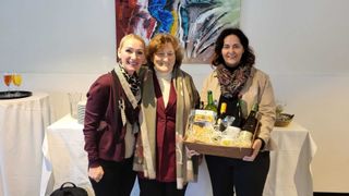 Three women are standing together, smiling, and holding a box filled with wine bottles and snacks. They are in front of a colorful painting on the wall.