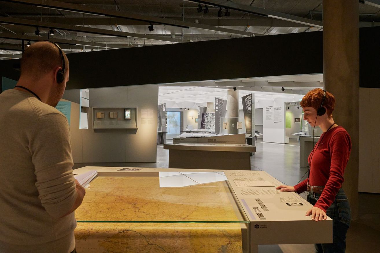 A man and a woman are in a museum. They are standing in front of a large map on a glass table. The woman is explaining something to the man. The room has many artifacts on display, including a large, curved black structure.