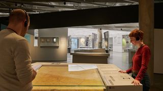 A man and a woman are in a museum. They are standing in front of a large map on a glass table. The woman is explaining something to the man. The room has many artifacts on display, including a large, curved black structure.