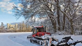 A red snowplow is clearing a path in a snowy field with trees and a building in the background.