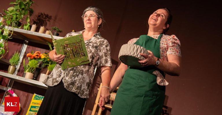 Two women in aprons stand together, one holding a book and the other a basket, with a stage backdrop featuring shelves with plants and flowers.