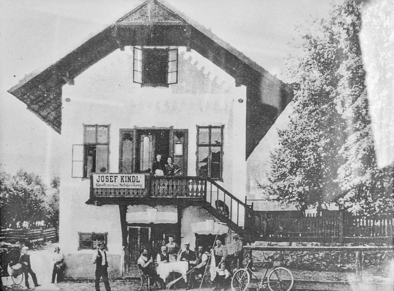 An old photo shows a group of people sitting around a table outside a house with a sign that reads Josef Kindl. They are dressed in old-fashioned clothing.