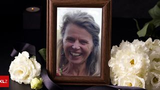 A smiling woman with gray hair in a brown frame surrounded by white flowers. A candle is visible on the left side.