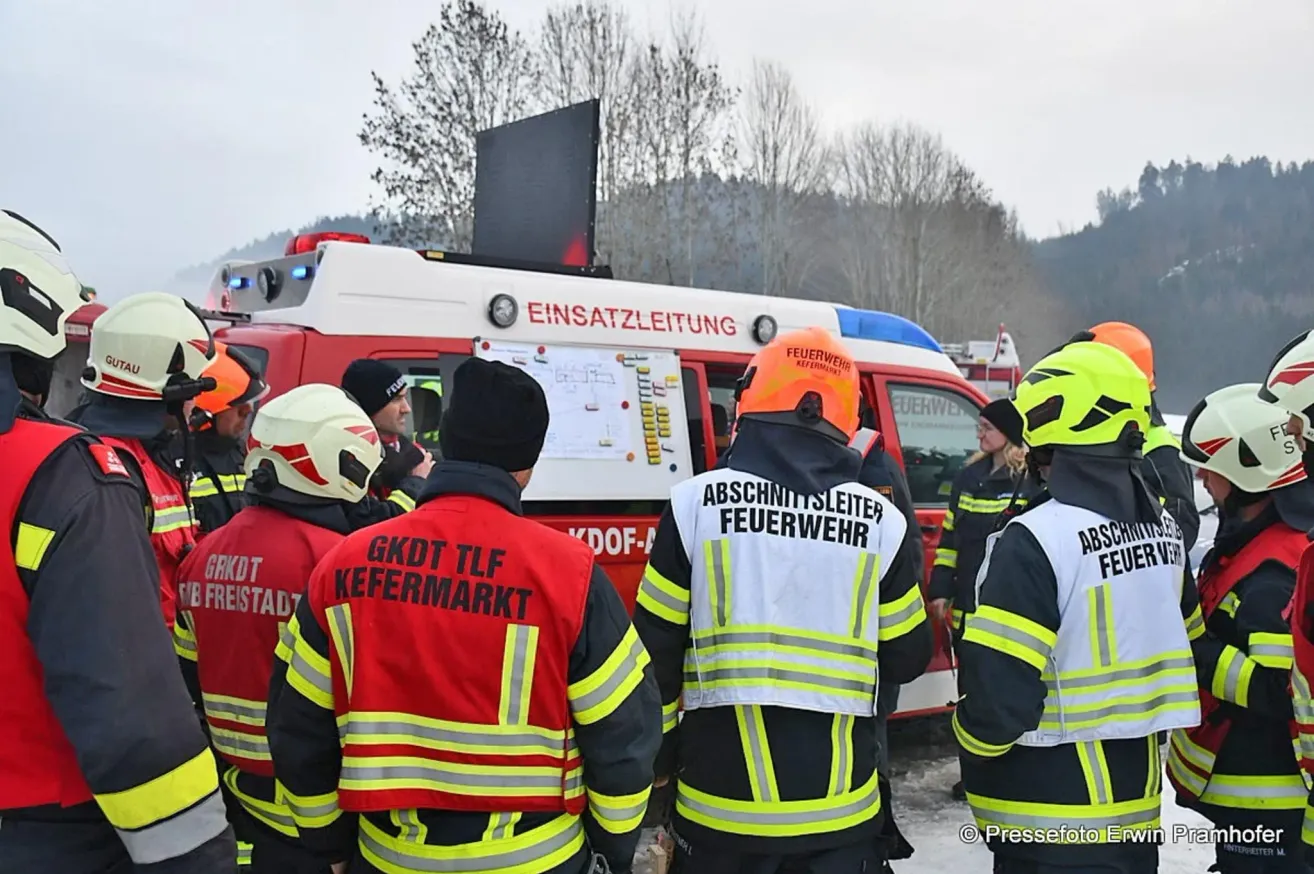 A group of firefighters stand in front of a vehicle. They wear uniforms with names and roles. The vehicle has red and white colors and displays the word Einsatzleitung.