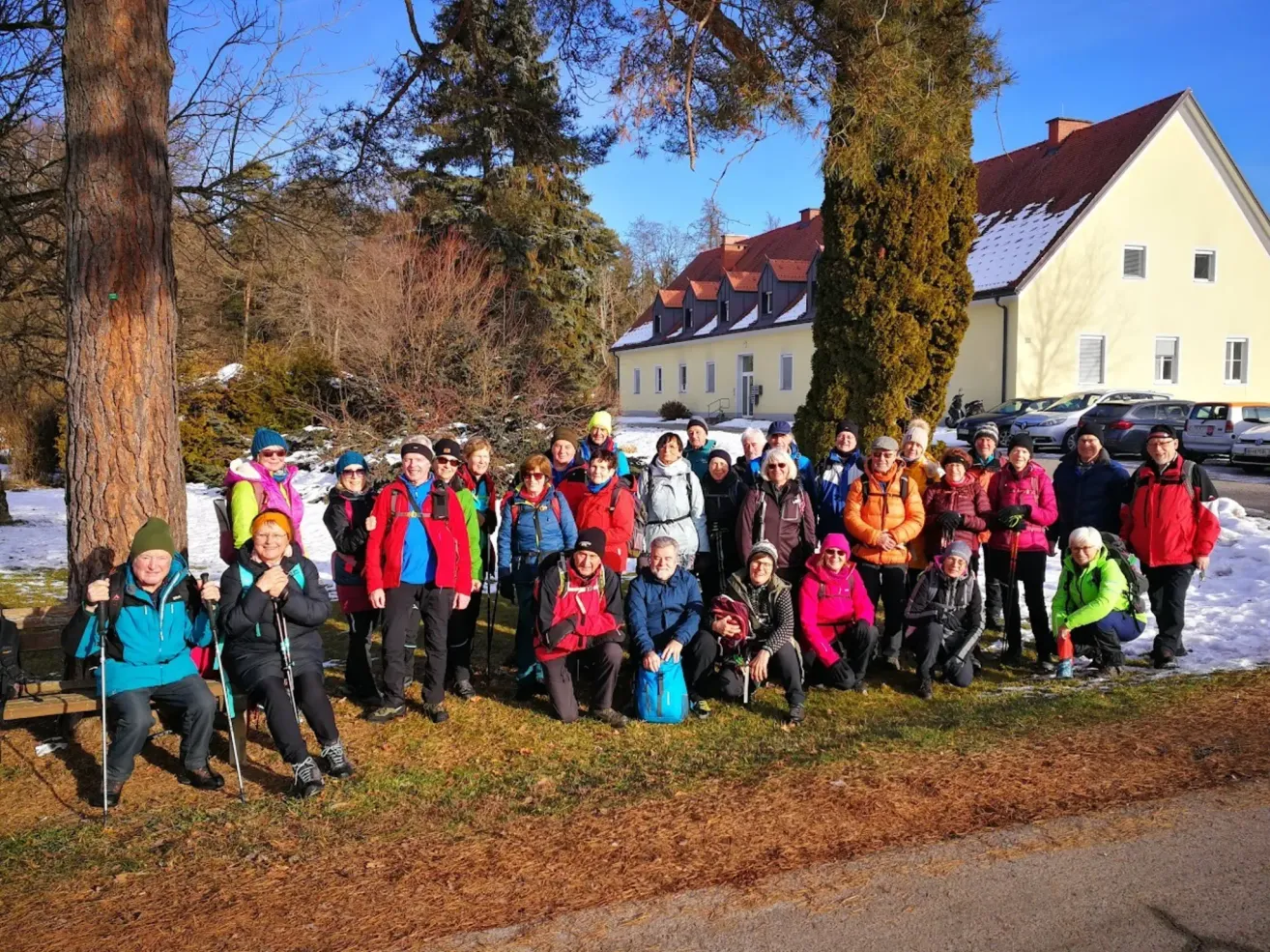 A group of hikers is gathered on a snowy path, wearing winter clothing, some with hats and sunglasses. A building with a red roof is in the background.