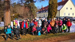 A group of hikers is gathered on a snowy path, wearing winter clothing, some with hats and sunglasses. A building with a red roof is in the background.