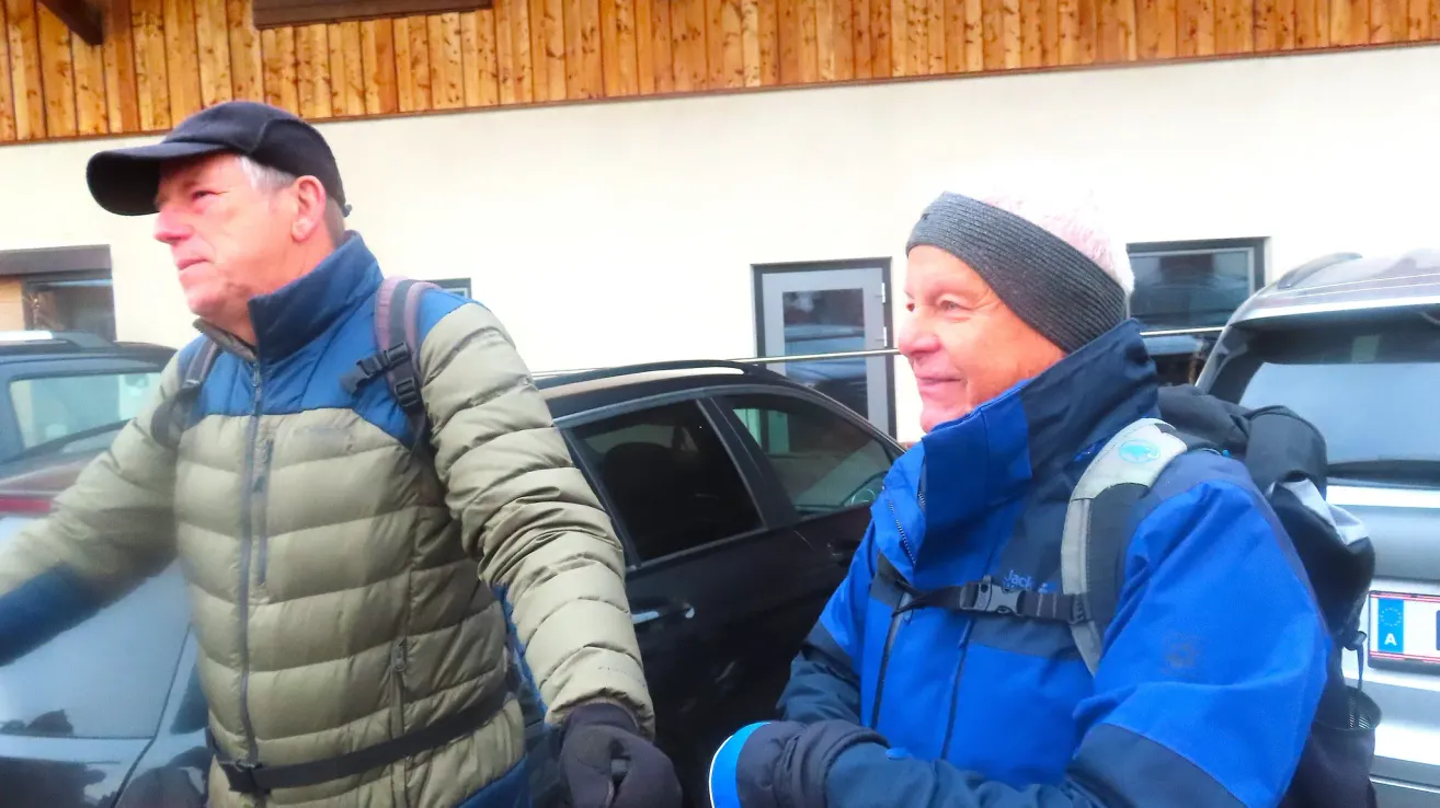 Two men in winter gear stand near a black car. One wears a backpack and black gloves, the other wears a beanie and blue jacket. They are outside with a wooden wall and a door behind them.