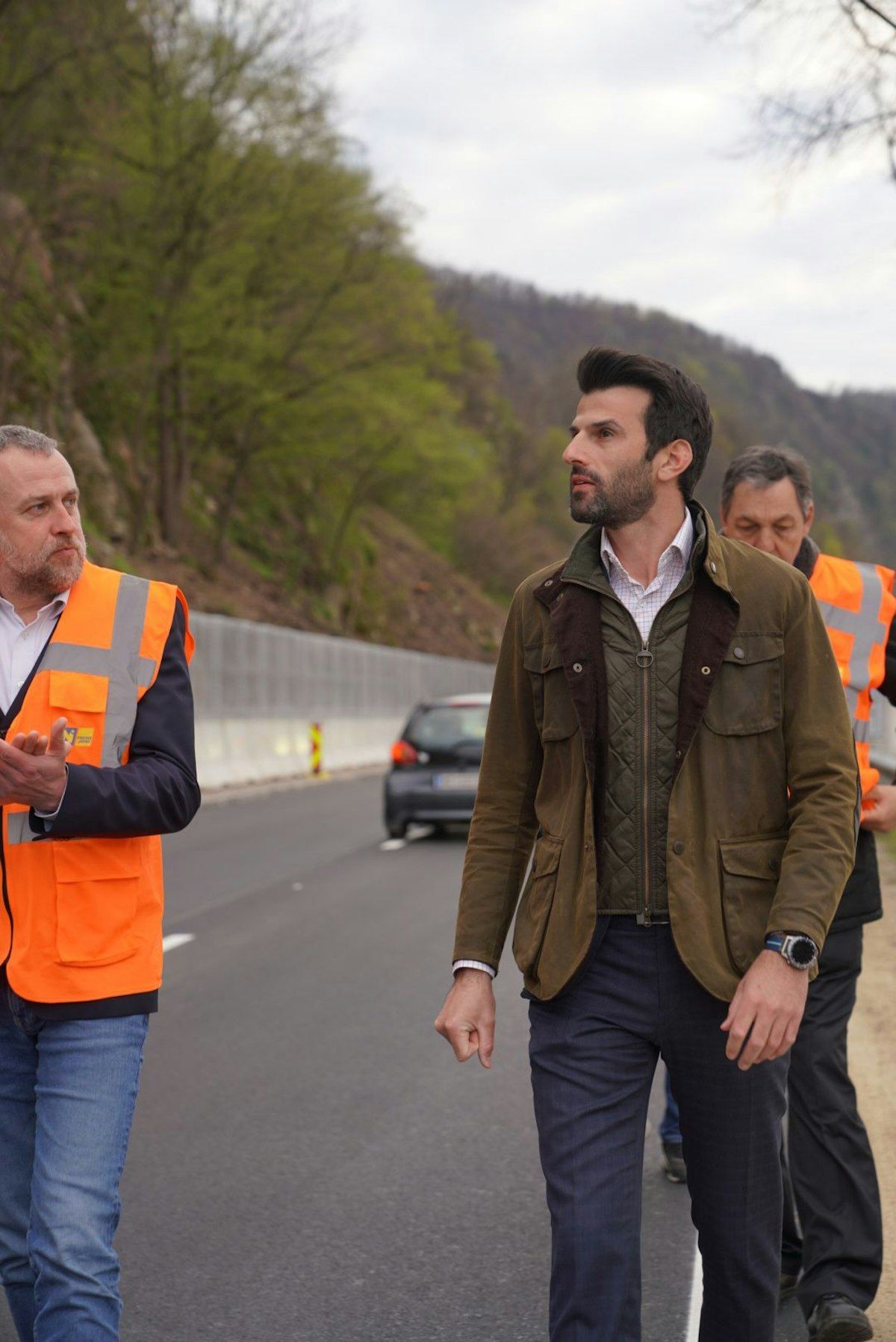 Three men walk on a road with a vehicle behind them. One man wears a reflective orange vest.