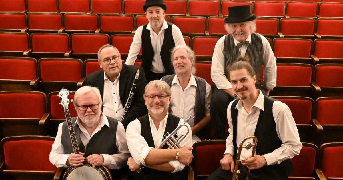 A band of musicians dressed in vests and hats are seated in a theater, each holding their respective instruments.