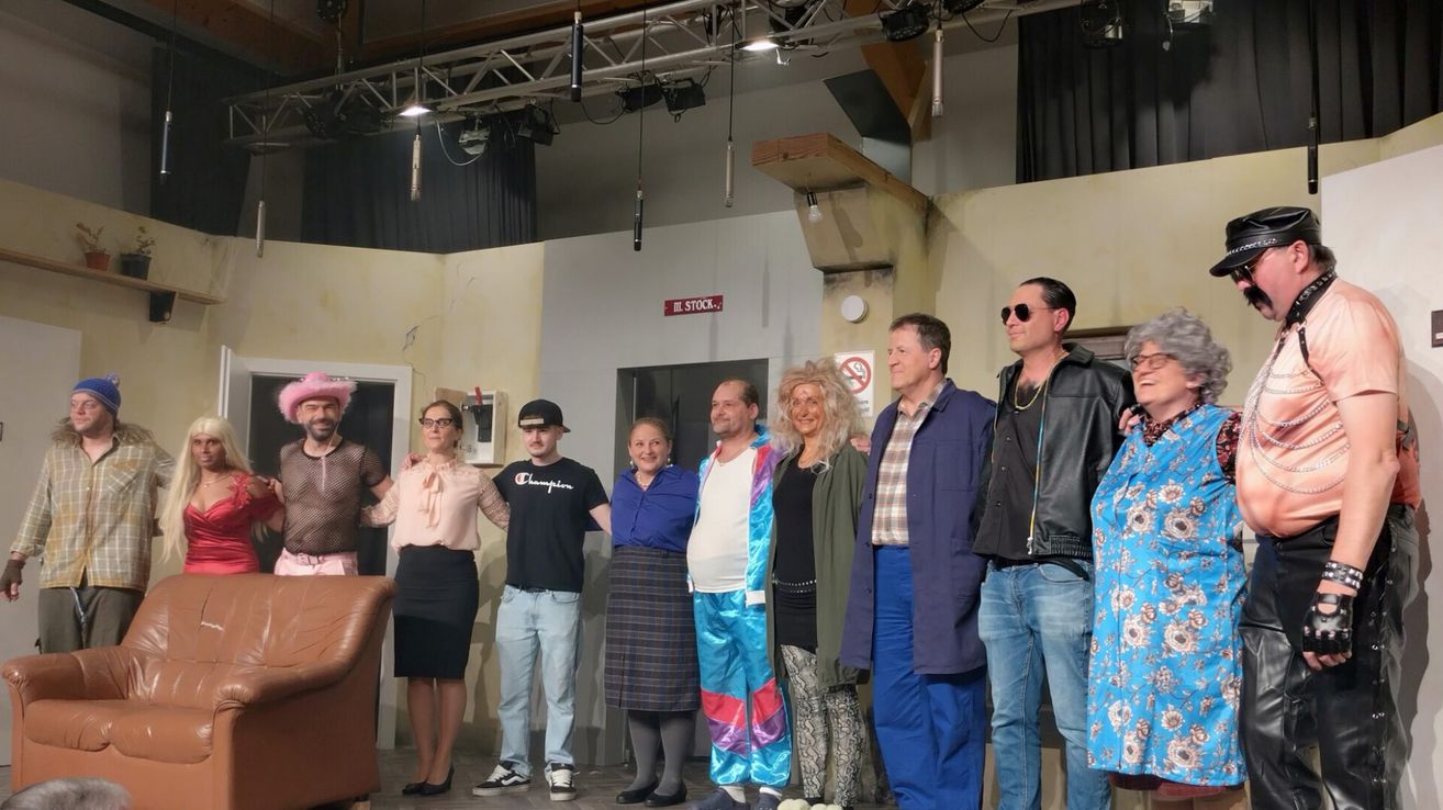 A group of people are standing together on a stage, possibly for a play. They are all dressed in costumes and some are wearing glasses. They are standing in a line, facing the front. Behind them is a wall with a door and a signboard. There are also some lamps hanging from the ceiling.