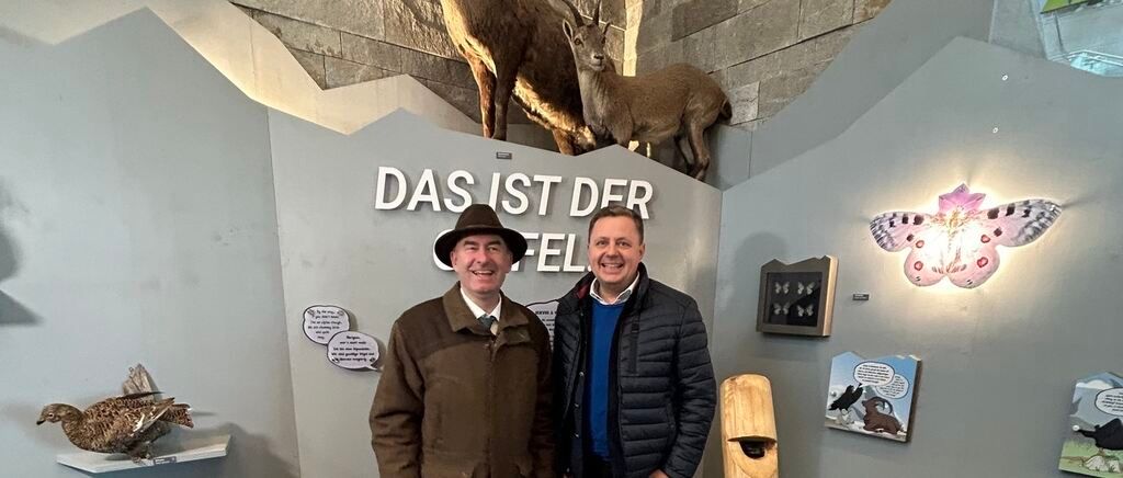 Two men are standing in front of a wall with animals mounted on it. The wall has a German text and some decorations.