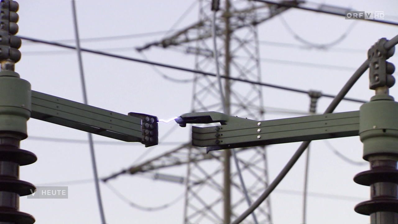 A close-up of two green electrical equipment parts connected by a sparking wire. In the background, tall power transmission towers and interconnected wires are visible.