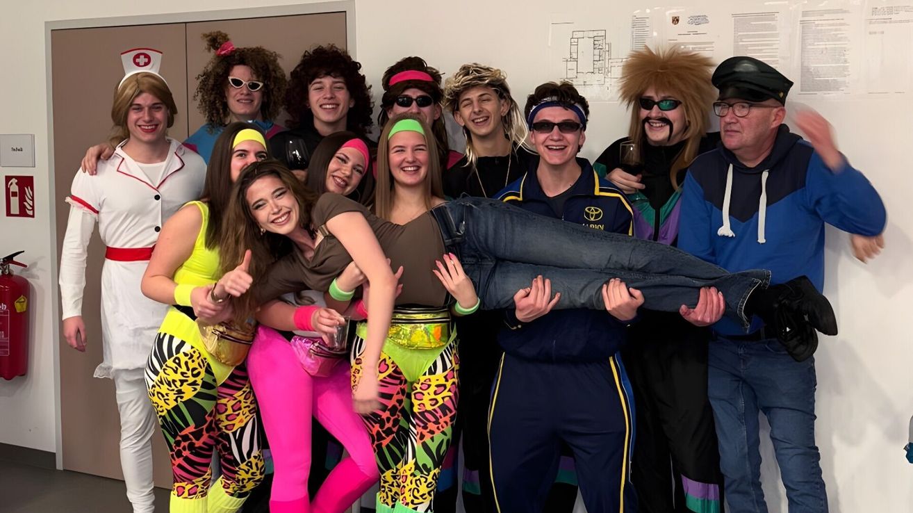 A group of young people in 80s-themed costumes pose for a photo. They are smiling and standing close together, with one person lying on the shoulders of others.