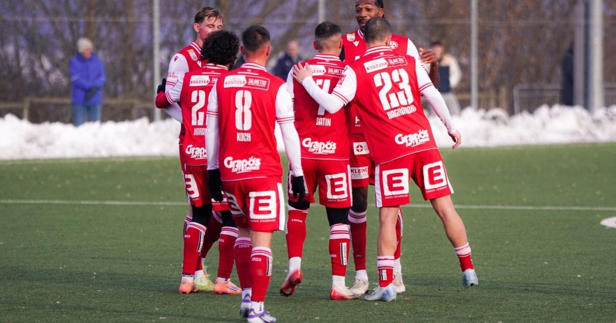 A group of soccer players in red uniforms celebrates on a snowy field. Players hug each other, numbers visible on their jerseys.