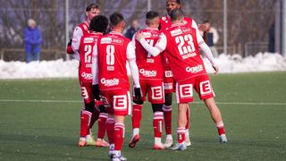 A group of soccer players in red uniforms celebrates on a snowy field. Players hug each other, numbers visible on their jerseys.