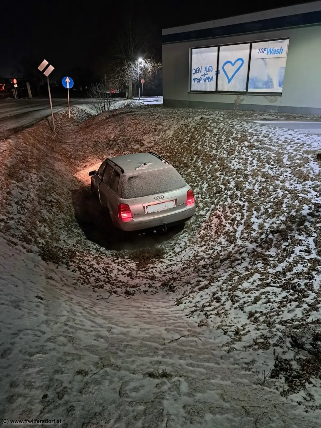 A car is stuck in a snowy ditch. It has license plate 000 and is covered in snow. A signboard with graffiti is nearby.