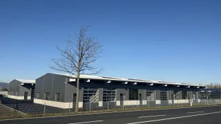 Ein großes Industriegebäude mit mehreren Fenstern und Solarpanels auf dem Dach befindet sich neben einer Straße mit weißen Markierungen und einem Zaun. Ein kahler Baum steht vor dem Gebäude.