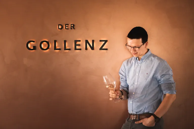 Ein Mann in einem blauen Hemd und mit Brille hält ein Weinglas vor einer Wand mit dem Wort Golenz.