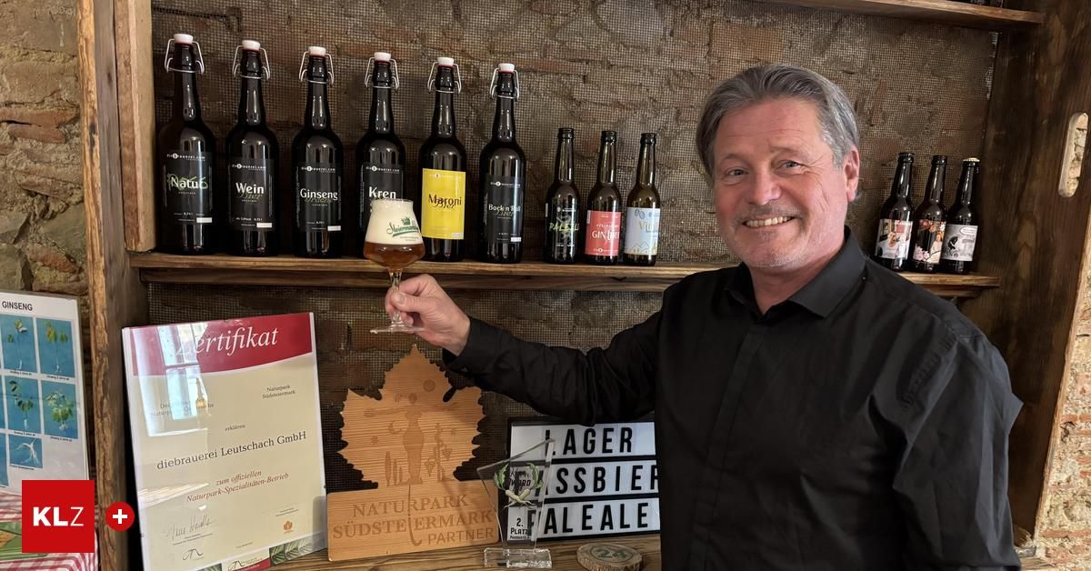 A man in a black shirt smiles while holding a wine glass, standing in front of a shelf with bottles and a wooden plaque.