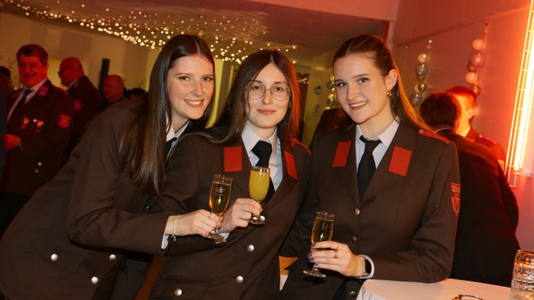 Three women in matching uniforms pose with wine glasses, smiling at the camera. They have red shoulder patches and are standing together in a festive setting.