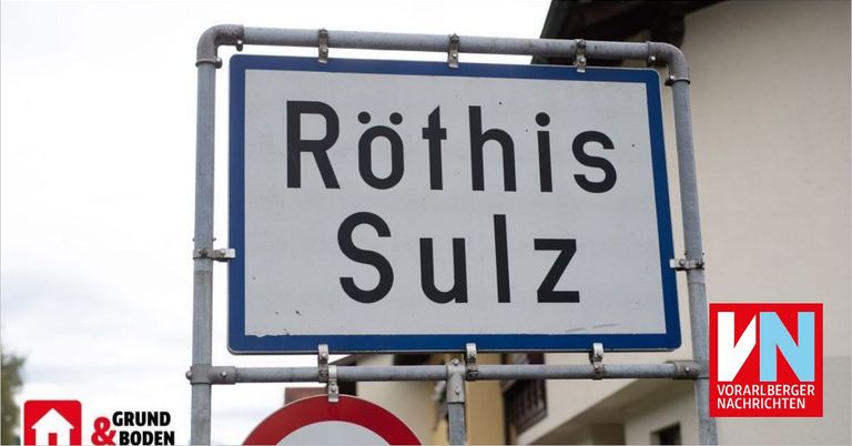 A white street sign with blue borders on a metal pole reads 'Rothis Sulz'. The sign is mounted above a red circular sign. Behind the sign is a building with multiple windows.