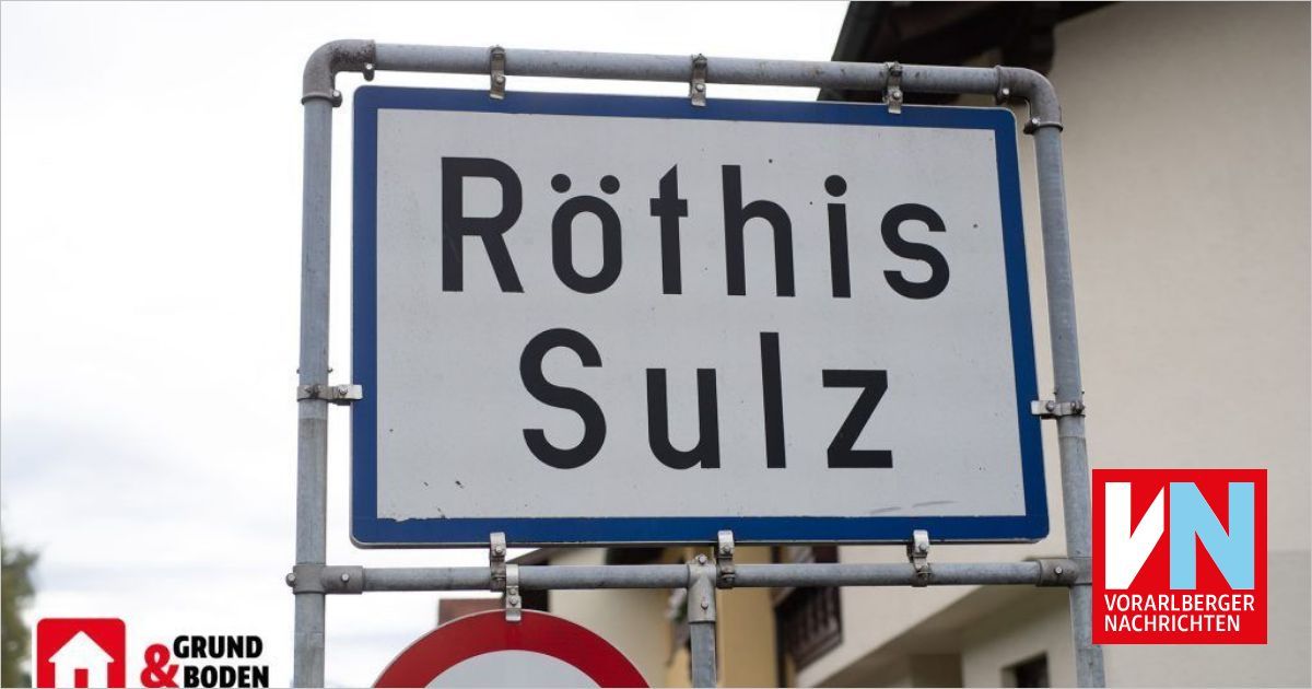 A white street sign with blue borders on a metal pole reads 'Rothis Sulz'. The sign is mounted above a red circular sign. Behind the sign is a building with multiple windows.