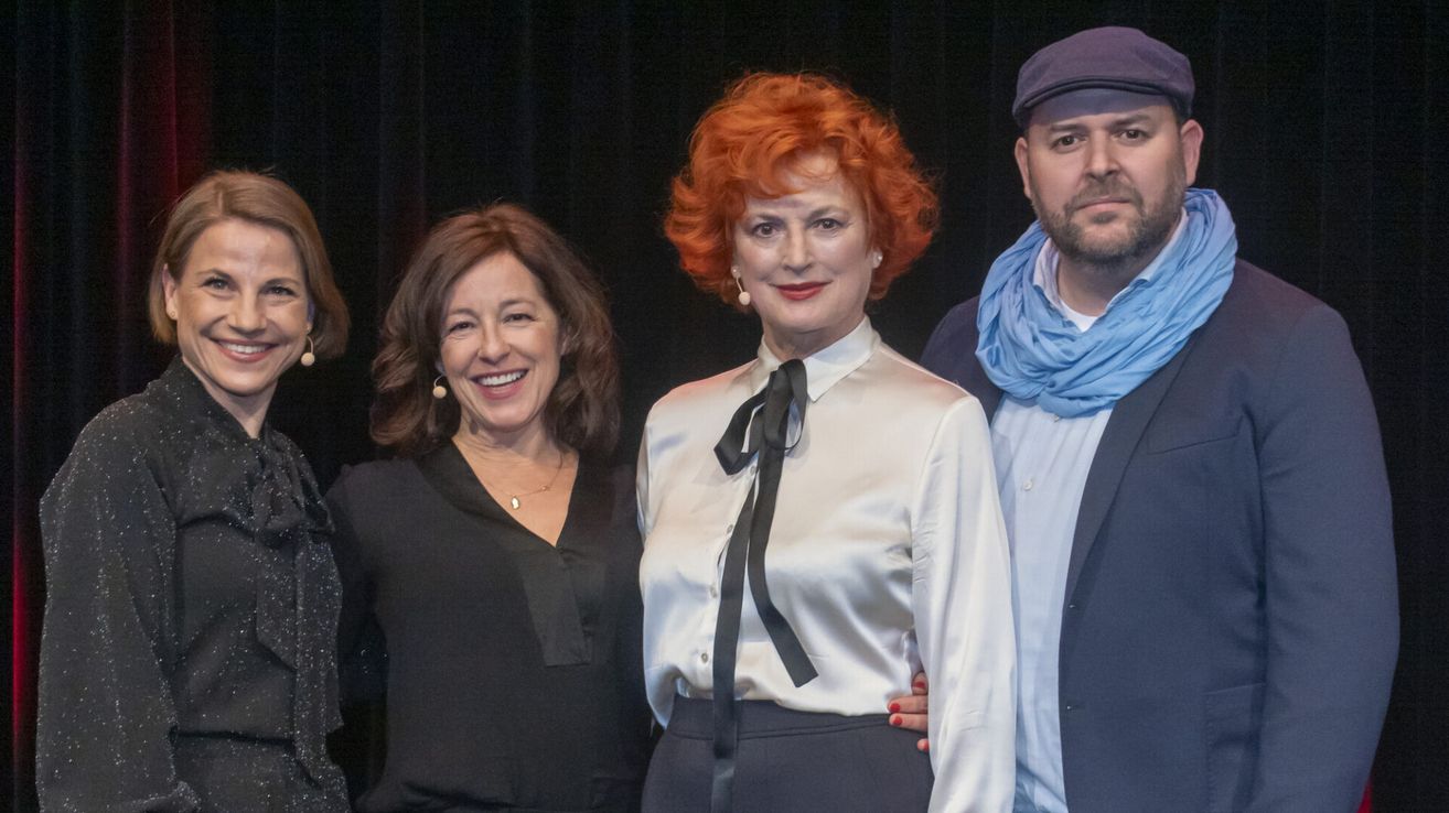Drei Personen posieren für ein Foto auf der Bühne, eine Frau mit roten Haaren, eine andere mit braunen Haaren und ein Mann mit einem blauen Schal. Sie alle lächeln.