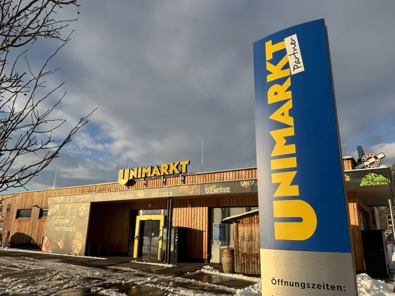 Die Vorderseite eines Unimarkt-Geschäfts. Ein Schild mit Unimarkt in gelben und blauen Buchstaben steht vor dem Eingang. Ein blaues und weißes Schild mit der Aufschrift 'Partner'. Schnee liegt auf dem Boden.