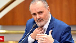 Ein Mann mit Bart und Schnurrbart, der einen blauen Anzug und ein weißes Hemd trägt, spricht in ein Mikrofon.