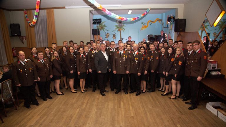 A group of people in uniform, likely military, stand together on a wooden floor. They are posing for a photo. A man in a suit stands between two men in uniform. The room has a blue wall with animal paintings.