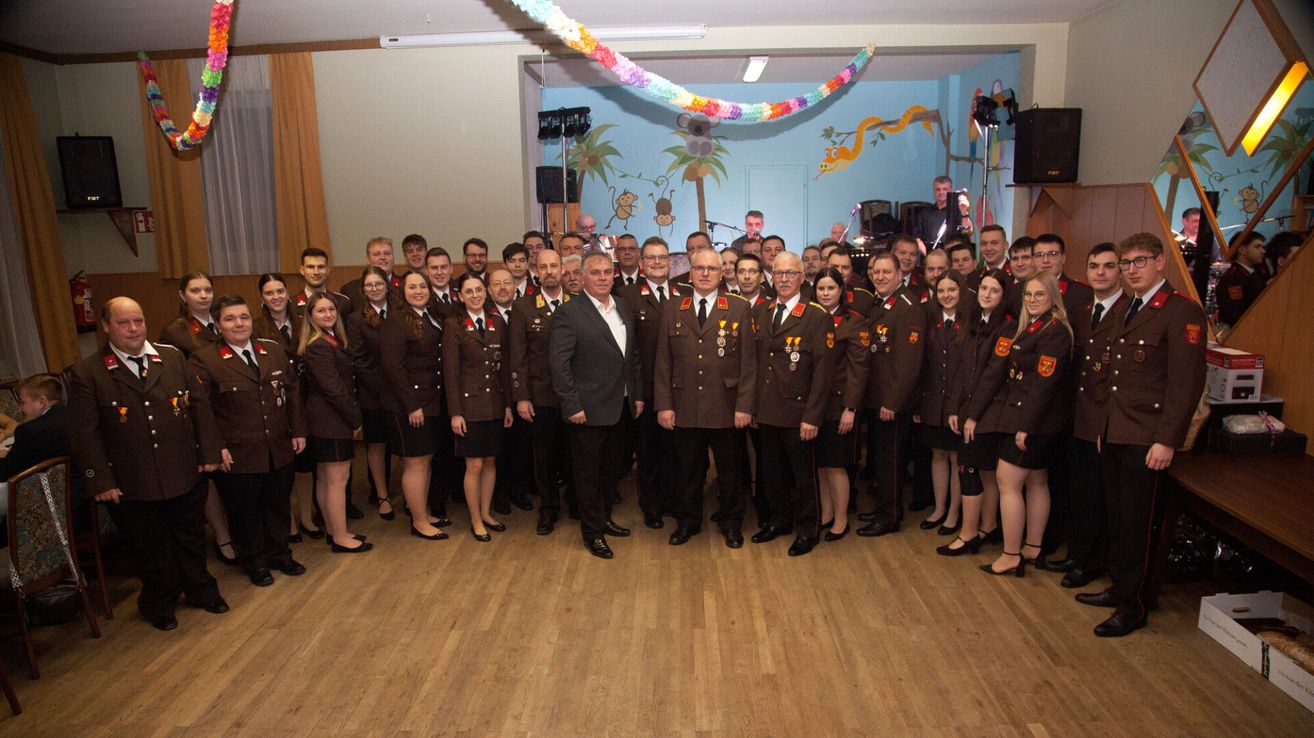 A group of people in uniform, likely military, stand together on a wooden floor. They are posing for a photo. A man in a suit stands between two men in uniform. The room has a blue wall with animal paintings.