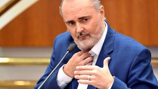 Ein Mann in einem blauen Anzug steht vor einem Mikrofon, er spricht und gestikuliert mit den Händen. Er trägt ein weißes Hemd und einen Bart.