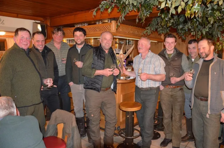 Eine Gruppe von Männern steht in einer rustikalen Bar, hält Weingläser und posiert für ein Foto. Der Mann in der Mitte hält eine Trophäe. Hinter ihnen steht eine Holzbar mit Regalen und einer Pflanze.