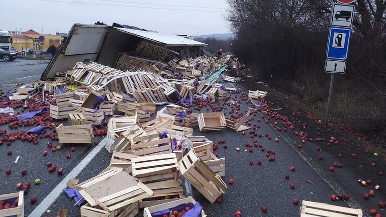 Ein Verkehrsunfall mit einem Lastwagen hat dazu geführt, dass mehrere Holzkisten ihren Inhalt, rote Äpfel, auf die Straße verschütten. Eine Person mit Helm steht in der Nähe des Wracks.