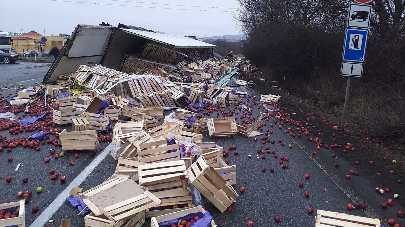 Ein Verkehrsunfall mit einem Lastwagen hat dazu geführt, dass mehrere Holzkisten ihren Inhalt, rote Äpfel, auf die Straße verschütten. Eine Person mit Helm steht in der Nähe des Wracks.