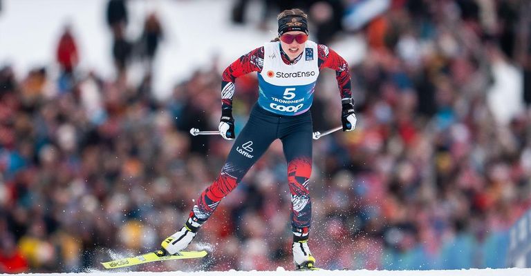 Eine Skifahrerin in Rot, Weiß und Blau fährt bergab und trägt eine Skibrille und Handschuhe. Im Hintergrund spritzt Schnee. Eine Menge schaut zu.