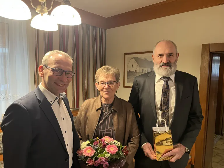 Drei Personen stehen in einem Raum zusammen. Der Mann auf der linken Seite hält einen Blumenstrauß, die Frau in der Mitte lächelt und der Mann auf der rechten Seite hält eine Geschenktüte. Ein Bilderrahmen hängt hinter ihnen an der Wand.