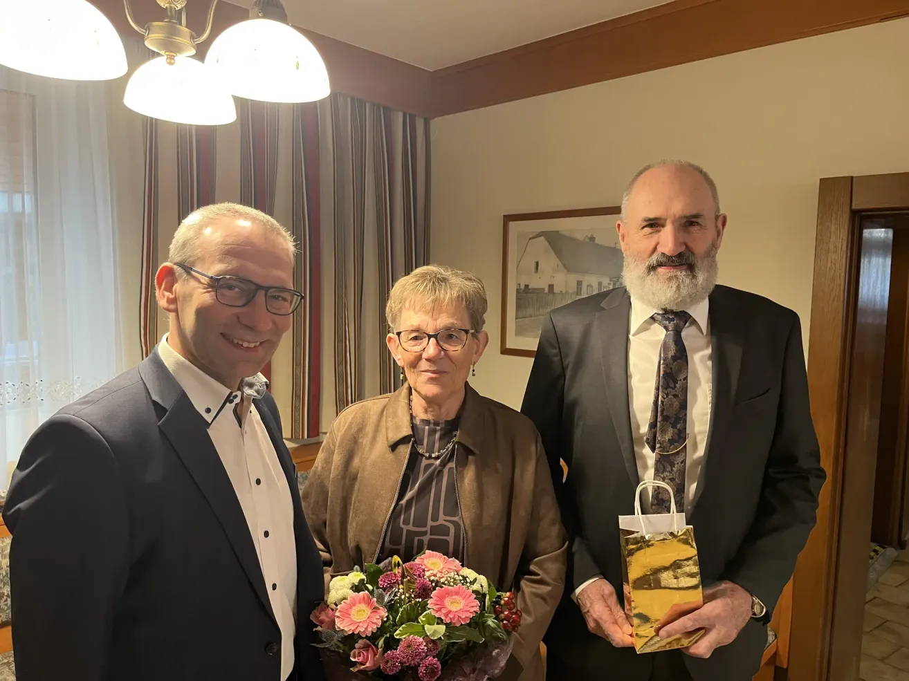 Drei Personen stehen in einem Raum zusammen. Der Mann auf der linken Seite hält einen Blumenstrauß, die Frau in der Mitte lächelt und der Mann auf der rechten Seite hält eine Geschenktüte. Ein Bilderrahmen hängt hinter ihnen an der Wand.