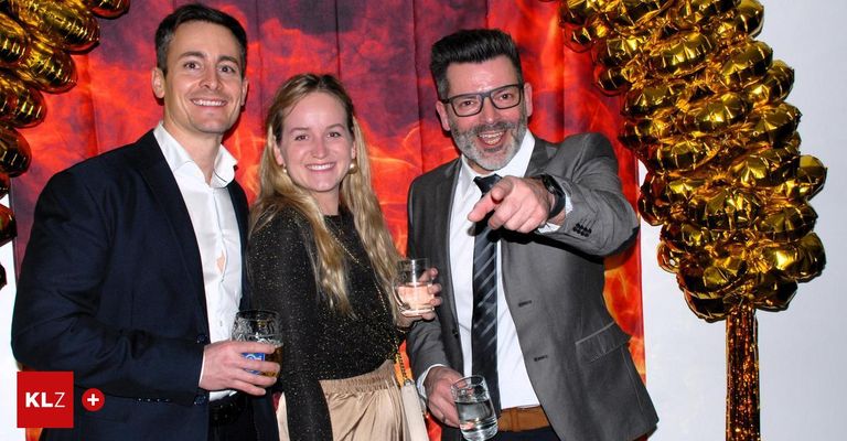 Drei Menschen stehen eng beieinander und lächeln für ein Foto auf einer Party. Ein Mann links hält ein Glas Bier, eine Frau in der Mitte hat ein Getränk und der Mann rechts zeigt. Hinter ihnen ist ein feuriger Hintergrund mit goldenen Dekorationen zu sehen.