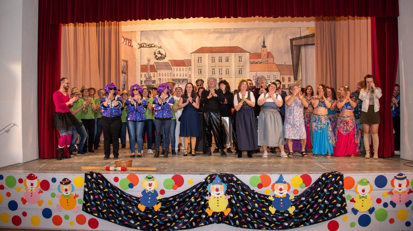 Eine Gruppe von Erwachsenen in verschiedenen Kostümen, einige mit Hüten, stehen auf der Bühne, klatschen und haben eine Kulisse mit Gebäuden im Hintergrund.