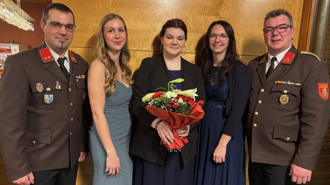 Drei Frauen und zwei Männer stehen eng beieinander für ein Foto, alle lächeln. Die Frau in der Mitte hält einen Blumenstrauß. Hinter ihnen ist eine Holzwand.