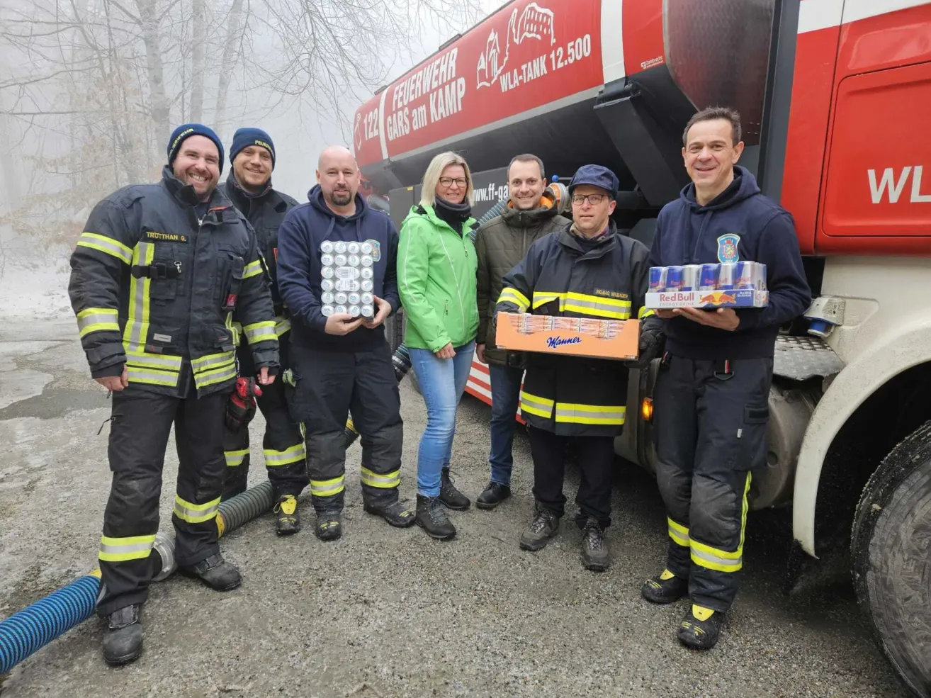 Eine Gruppe von Feuerwehrleuten in Uniformen, einer hält eine Dose Red Bull und der andere eine Box Manne, posiert vor einem Feuerwehrauto mit einem großen Tank.