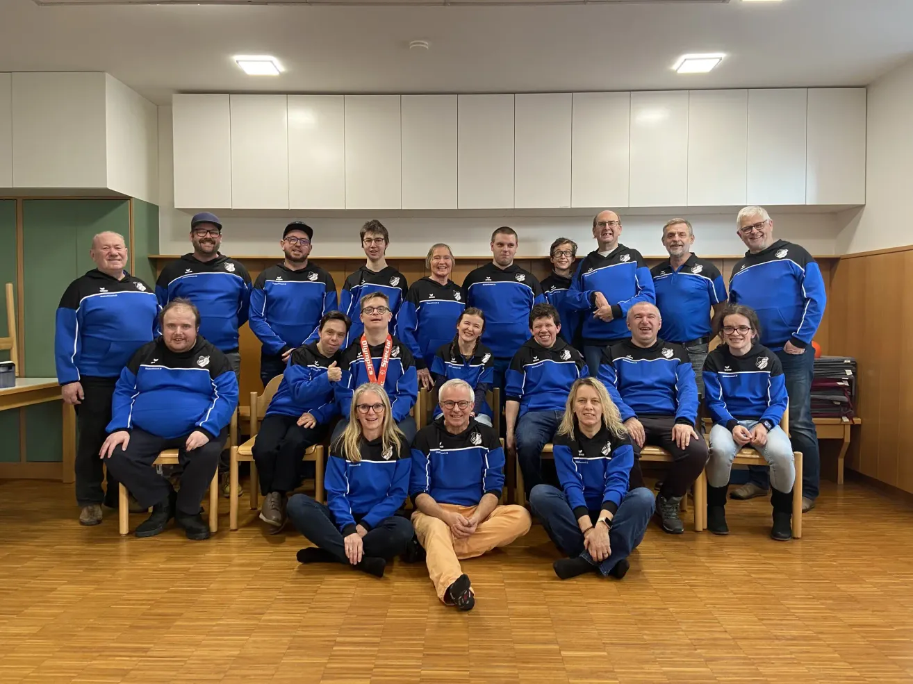 Ein Team in blauen Jacken posiert für ein Foto, einige sitzen auf Stühlen und andere stehen, in einem Raum mit Holzböden und weißen Schränken.