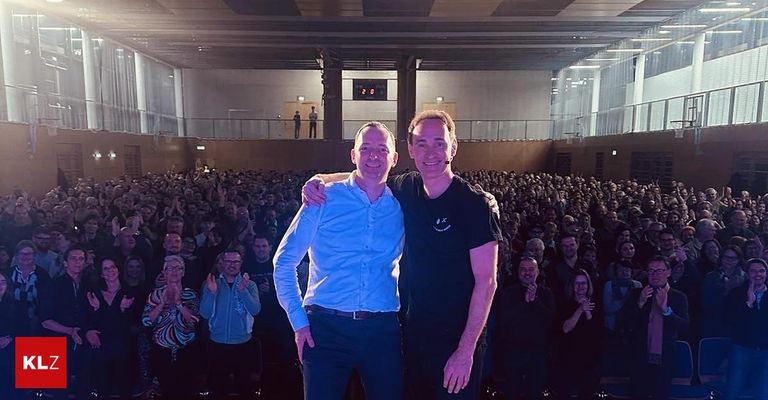 Zwei Männer stehen nebeneinander vor einer jubelnden Menge in einem Theater, einer in einem blauen Hemd und der andere in einem schwarzen T-Shirt.