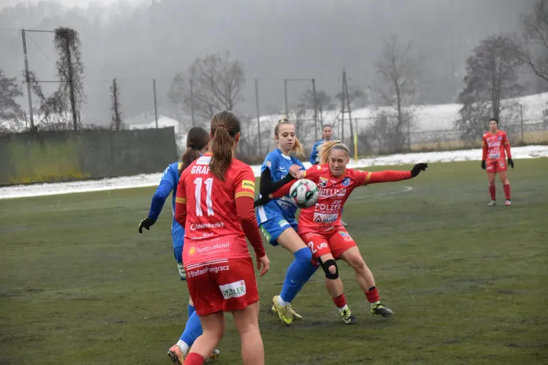 Fünf Fußballerinnen spielen auf einem Rasenfeld. Eine Spielerin in Rot, mit der Nummer 11, versucht den Ball zu schießen. Eine andere Spielerin in Blau versucht sie zu blockieren.