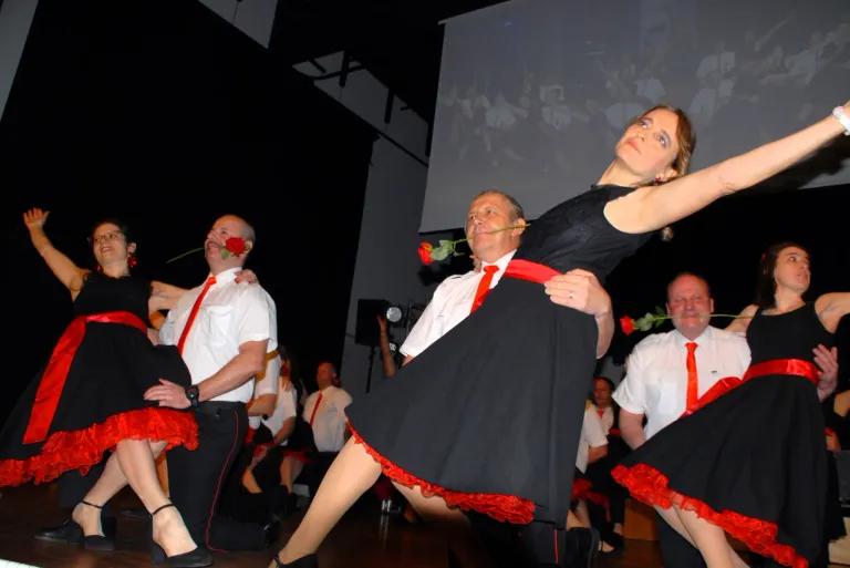 Eine Gruppe von Menschen, die in Schwarz und Rot gekleidet sind, tanzt auf einer Bühne mit einer Projektionsleinwand dahinter. Zwei Männer halten Rosen.