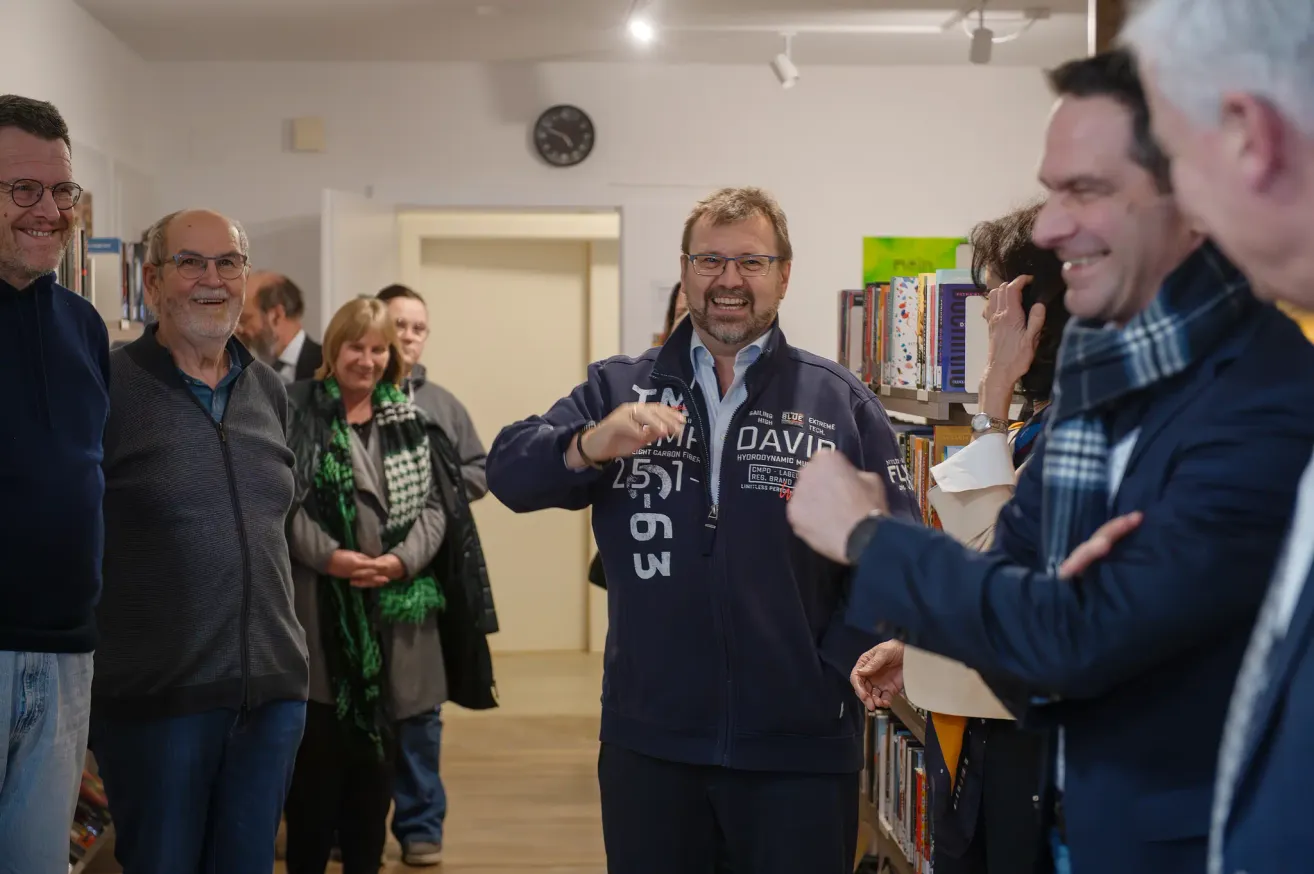 Eine Gruppe von Menschen in einem Raum, einer trägt eine blaue Jacke mit der Nummer 257, lächelt und unterhält sich mit den anderen.