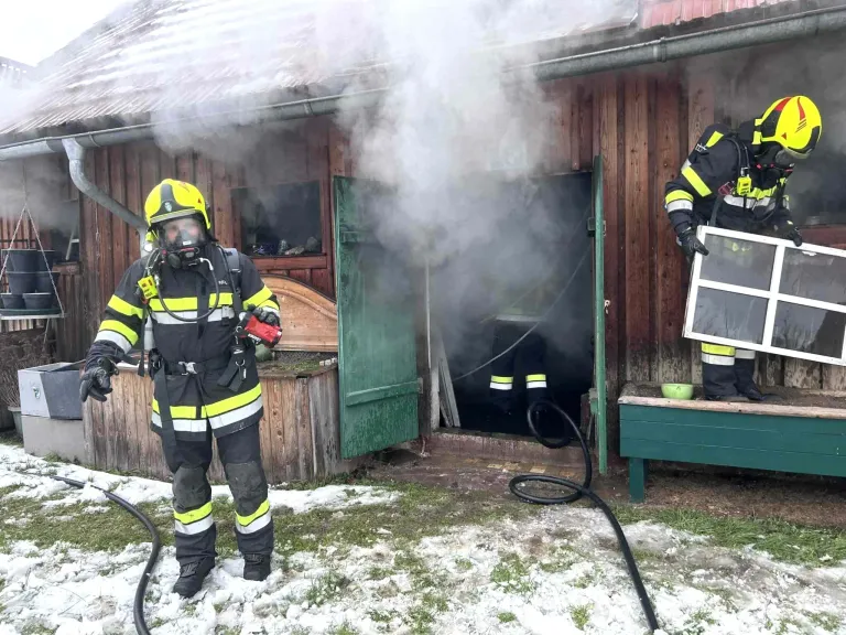 Feuerwehrleute in voller Ausrüstung stehen vor einem Holzgebäude, aus dem Rauch austritt. Ein Feuerwehrmann hält einen Feuerlöscher, während ein anderer in der Nähe einer offenen Tür steht.